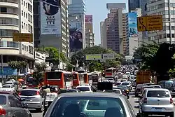 Looking down a busy road, which is banked on both sides by tall buildings, some of which are covered in advertisement billboards