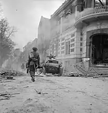 A man walks away from the camera, down a street littered with debris. Ahead of him is a small armoured vehicle.