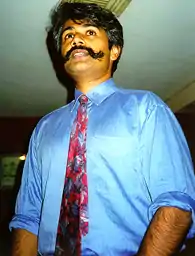 Photo of Tunku Varadarajan, dressed in blue shirt, sleeves rolled up, with red decorative necktie. He's sporting a handlebar mustache.