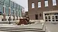 A statue consisting of a large, bronze "M", with a gold Goldy Gopher leaning on it, located on a slightly raised platform. Behind and to the right is a brick wall with a window and several doors. In the background to the left and behind several metal bike racks is a building with a brick, concrete, and glass facade.