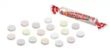 An image of Smarties tablet candies on a white background. On the right is a roll of candies consisting of 15 multicolored tablets in a clear film wrapper. The wrapper displays the Smarties Candy Company logo. On the left are a selection of loose Smarties candies.