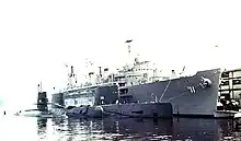 Three submarines are moored side by side to a large surface ship, which in turn is held to a dock in the background.