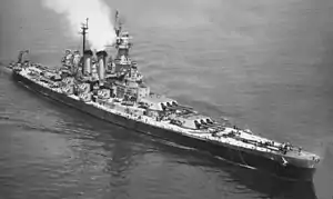 A large dark gray warship is underway at sea, with steam coming from the smokestacks