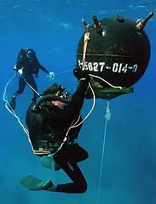 A diver appears to work on a large spherical mine, with another diver observing from a distance in the background