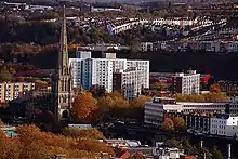 A view looking down towards a tall church spire which rises high above the surrounding trees and large modern buildings, with rows of hillside housing in the distance