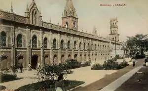 A panoramic depiction of a two-story nineteenth-century building alongside a square with several small trees.