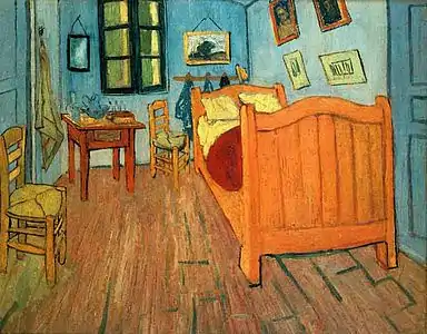  A narrow bedroom with wooden floor, green walls, a large bed to the right, a 2 straw chairs to the left, and a small table, a mirror and a shuttered window on the back wall. Hanging over the bed are several small pictures