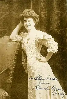 A white woman, standing, leaning against a chair; her hair is in a bouffant updo, and she is wearing a light-colored dress with a prominent lace overlay.