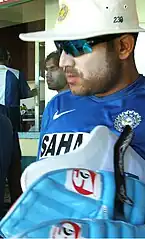 A man in the blue Indian cricket practice kit, wearing sun cream, sunglasses and a hat carrying his batting pads. Others can be seen in the background.