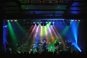 Members of a band, long-haired and dressed in black, perform under multi-colored lights.