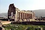 A picture of a rectangular building whose back side has been demolished. A green mountain range fills the background.