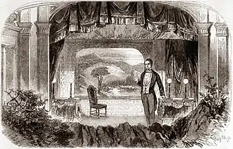 White, middle aged man in 19th-century evening clothes on a small stage with a countryside scene as the backdrop