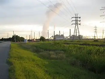 Photo shows a power plant with four smoke stacks. A train sits on a nearby railroad track.