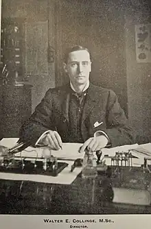 Walter Collinge in a suit, sat a table covered with laboratory equipment