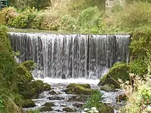 A weir with a river cutting a rough winding channel roughly 2 metres deep flowing through a wooded valley with paths.