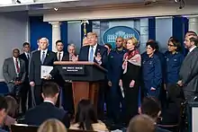 Trump speaks in the West Wing briefing room with various officials standing behind him, all in formal attire and without face masks