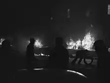 People silhouetted against cars in the background, which are engulfed in flames. One person is bent over and holding a doughnut-shaped object.