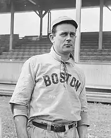 A man in a white cap and baseball jersey with a high collar and "Boston" written across the chest stands in front of a grandstand.