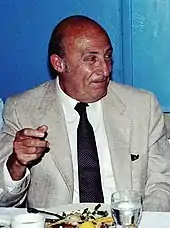 A photograph of an elderly man, seated and wearing a light-gray suit and tie.