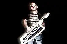 Stylized image of Yinon Yahel with black background, wearing a black t-shirt with white lettering, ripped blue jeans, and white plastic frame sunglasses, playing a portable keyboard strapped over his shoulder