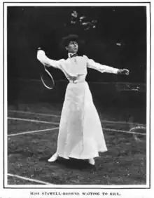 A white woman standing on a tennis court in tennis whites, including an ankle-length skirt, holding a tennis racquet behind her head in a position of readiness